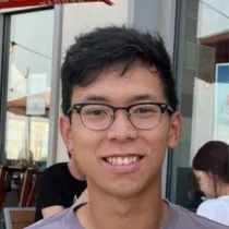 Young man with glasses smiling indoors at a café with other people in the background.