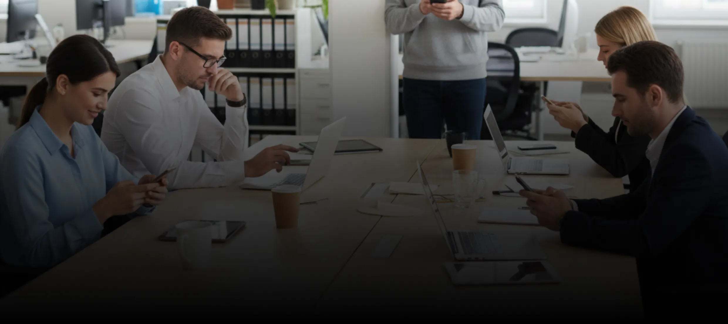Four colleagues sitting at a table in an office, using laptops and smartphones, with one person standing and holding a phone in the background.