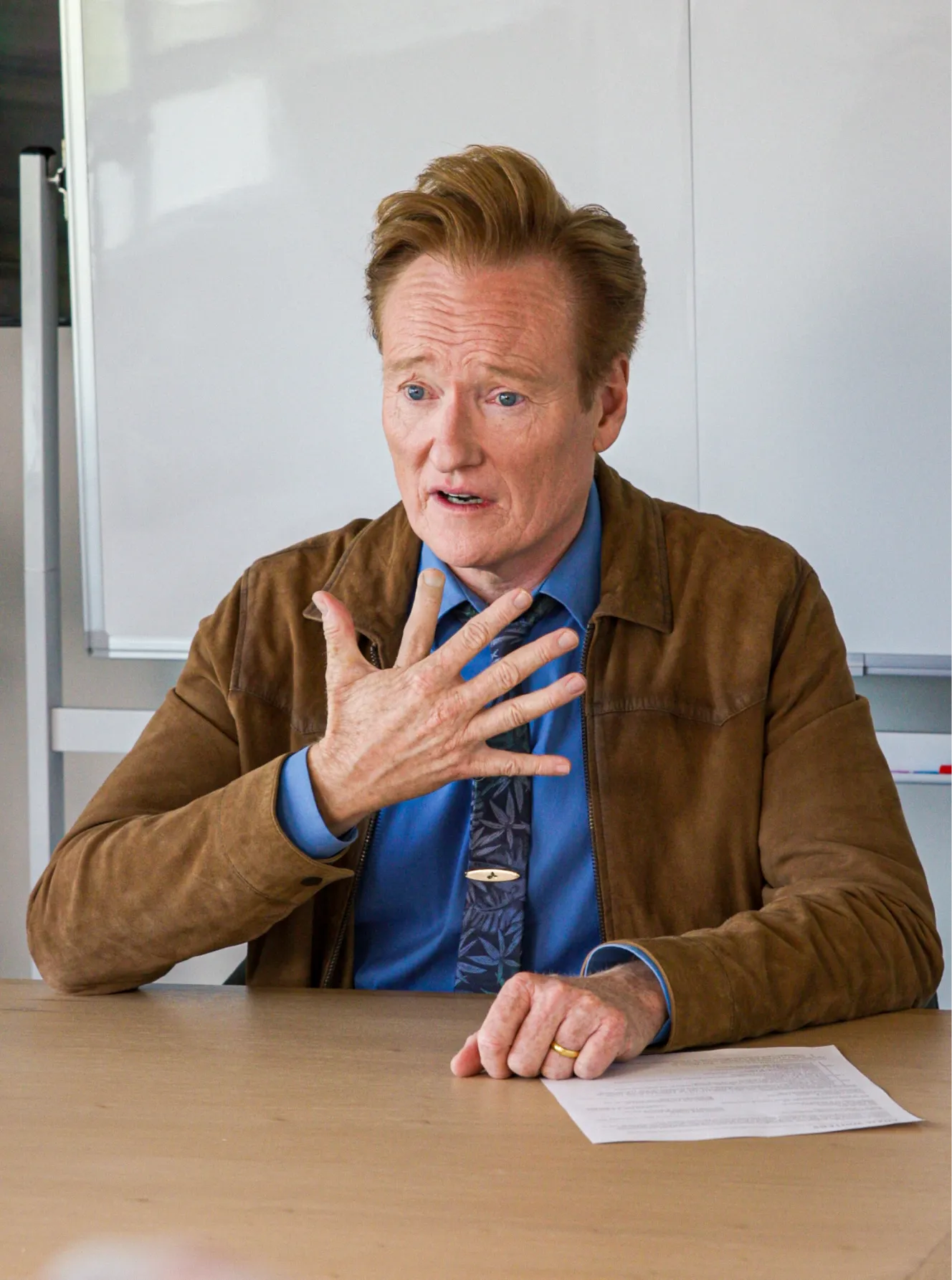 Man in brown jacket and blue shirt sitting at a table with paper, gesturing with open hand.
