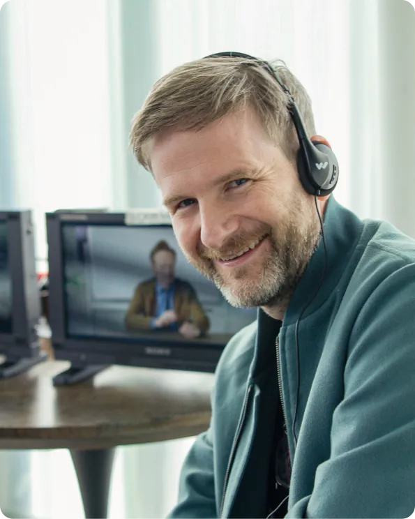 Smiling man wearing headset in front of a monitor showing a person in a video call.