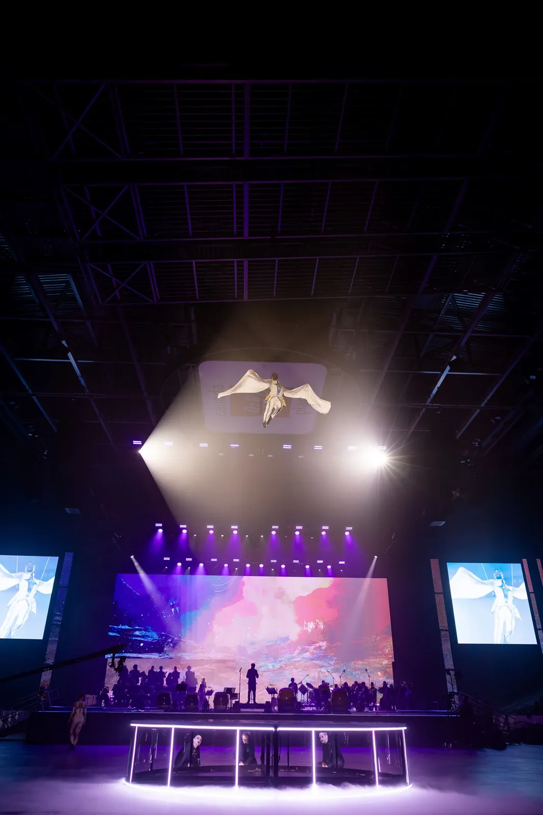 Aerial harness performer flying above an arena stage under spotlights