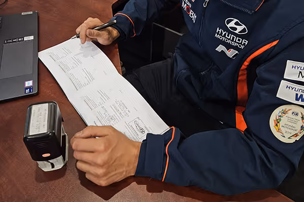 Mechanic wearing a Hyundai Motorsports jacket stamping and signing a Hyundai vehicle logbook on a wooden table next to a laptop.