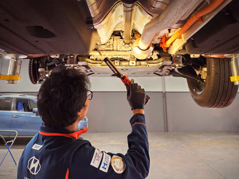 Mechanic inspecting the underside of a raised car using a handheld light in a garage.