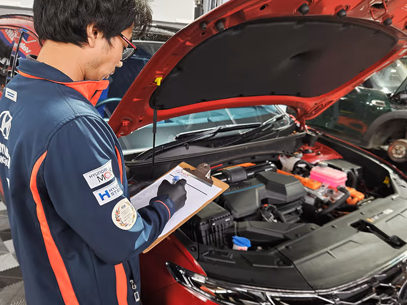Mechanic inspects and checks a red Hyundai Tuscon’s engine with a clipboard in hand.