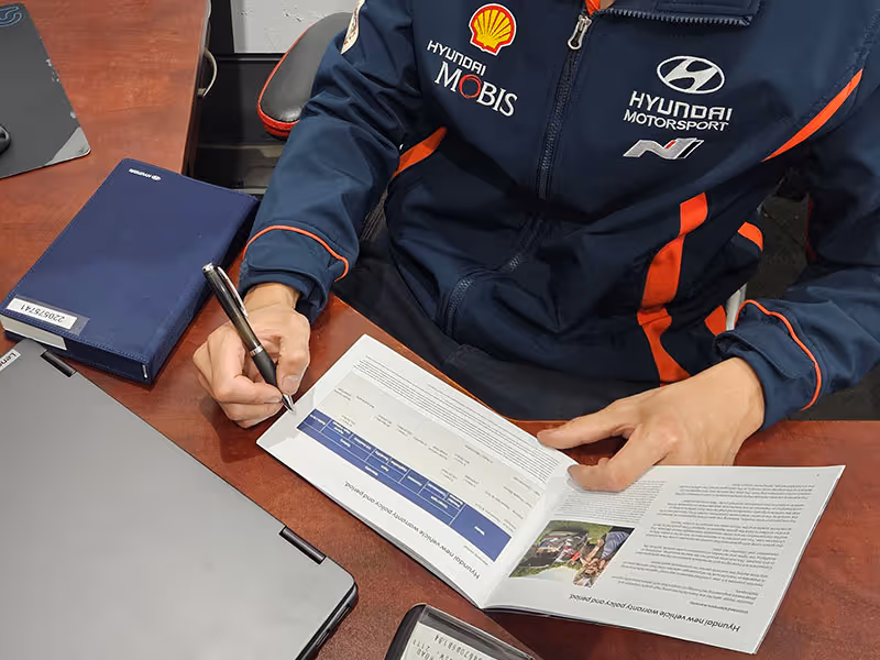 Mechanic in Hyundai Motorsport jacket reviewing and checking a Hyundai warranty booklet on a desk.