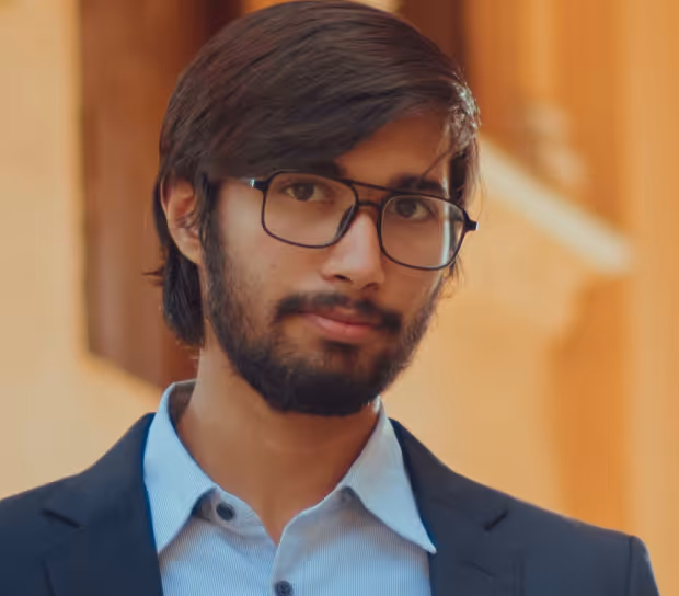 Young man with glasses and a beard wearing a blue dress shirt and dark blazer, standing outdoors.