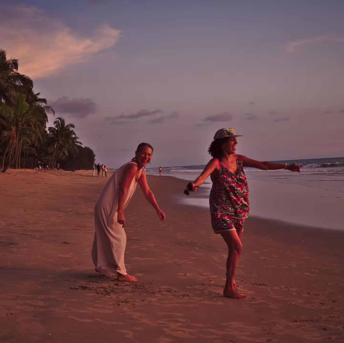 Two women enjoy an active, carefree walk along a tropical beach at sunset, surrounded by palm trees and gentle ocean waves, capturing the wellness and relaxation experience of an Ayurooms retreat by the sea.