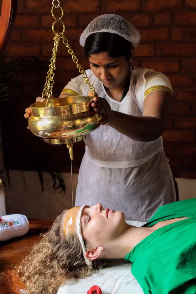 Ayurvedic therapist performing Shirodhara therapy, gently pouring a stream of warm oil onto a guest’s forehead at an Ayurooms wellness retreat in India.


