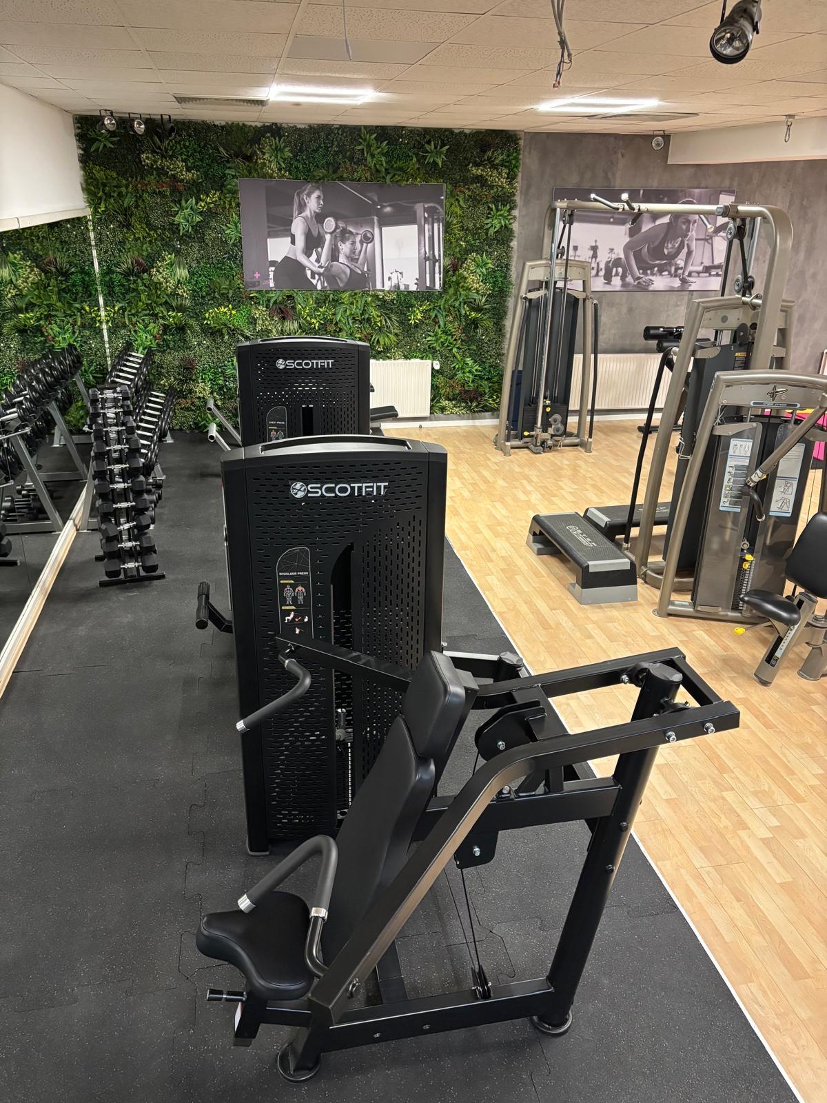 Modern gym with Scotfit weight machines, dumbbells on racks, and green plant wall and workout photos.