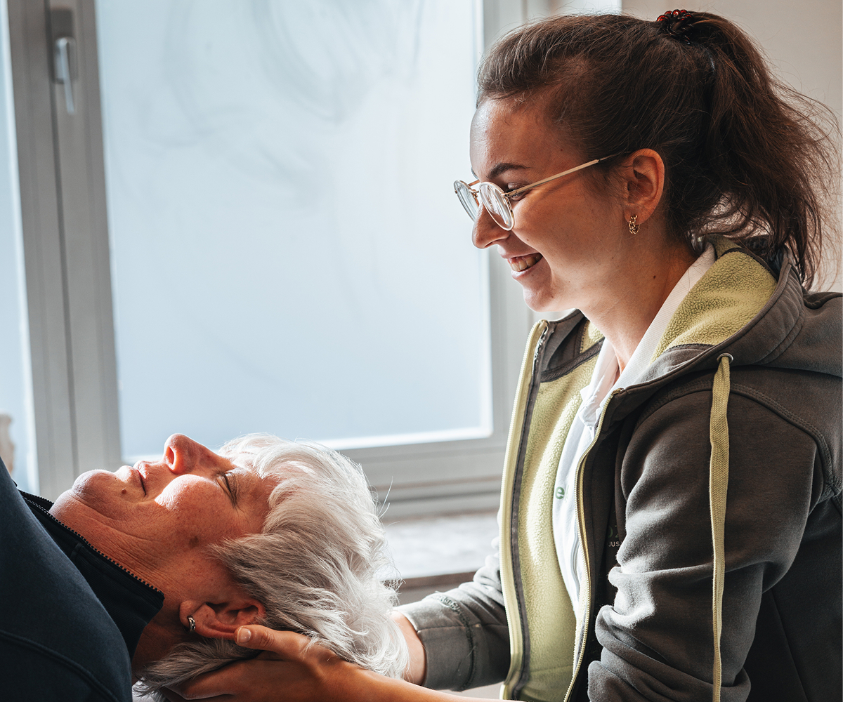 Smiling woman with glasses holding the head of an older woman lying down by a window.