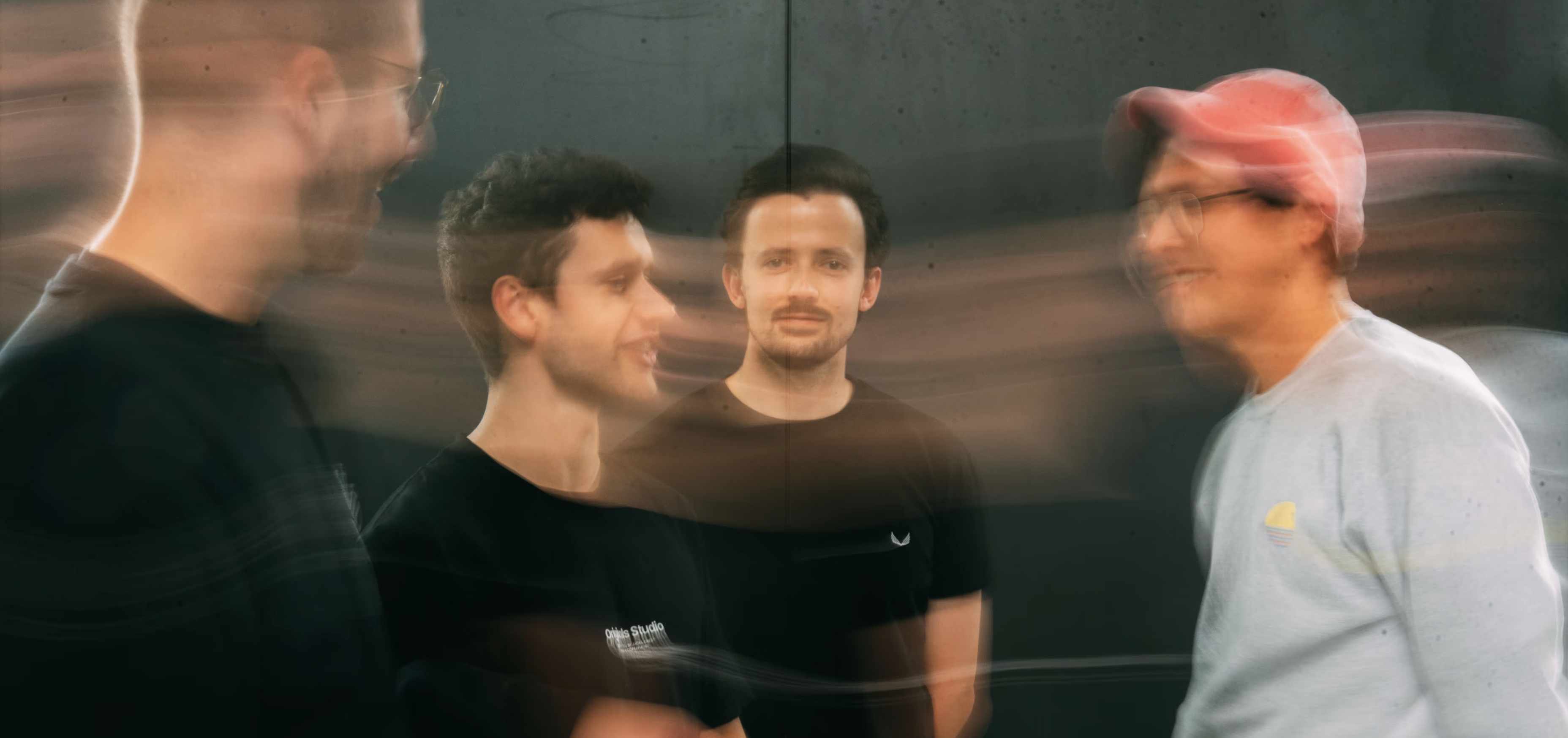 Vier jonge mannen, waarvan drie bewegingsonscherp en één duidelijk in het midden, staand tegen een donkere achtergrond.