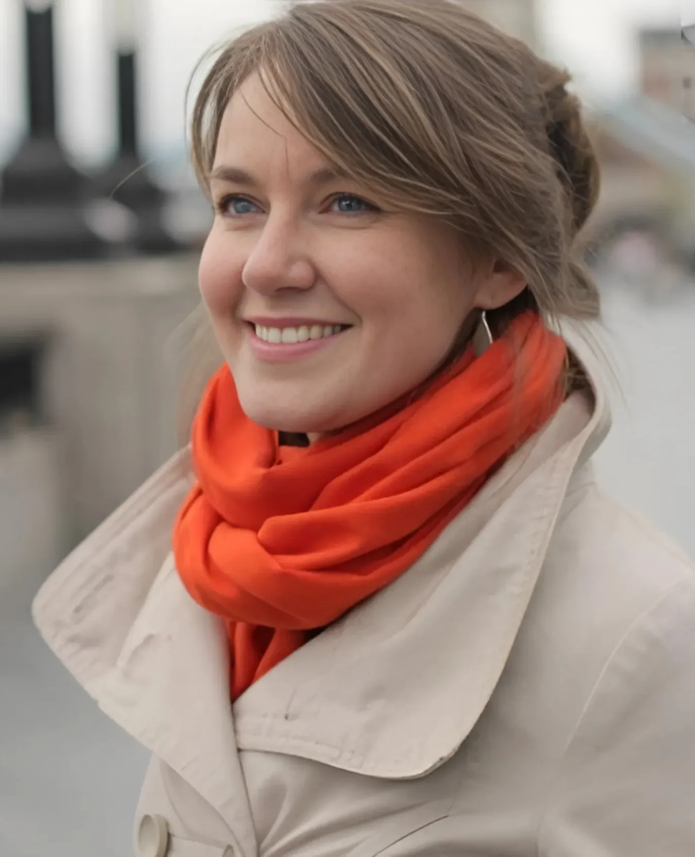 Arune, Doctor at Pilėnų klinika, smiling outdoors, light brown hair tied back, wearing a beige coat and bright orange scarf.