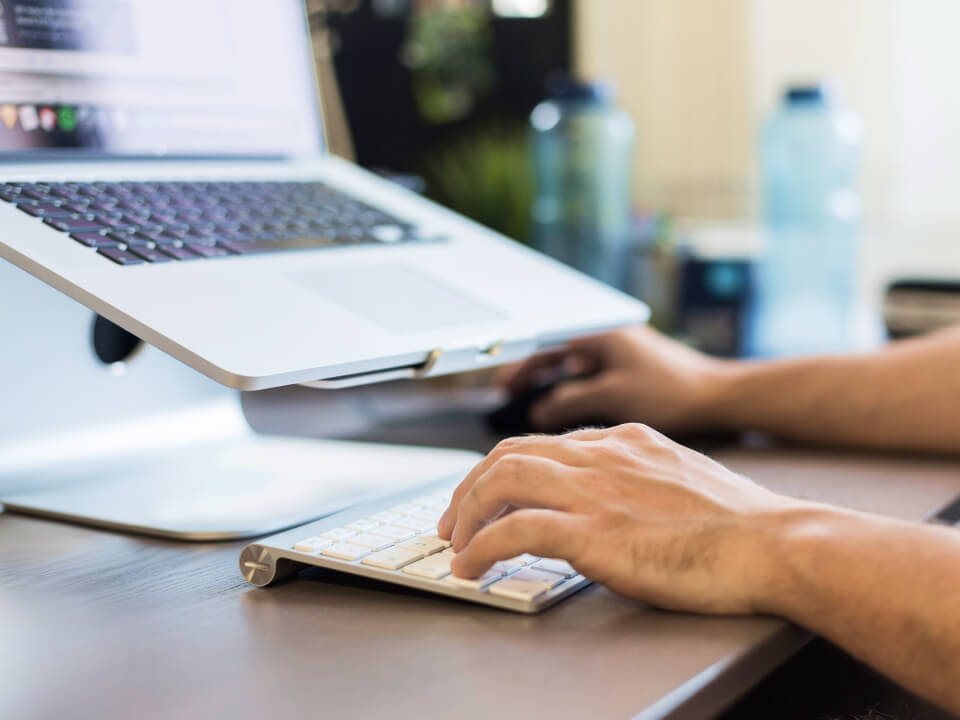 Das Bild zeigt eine Nahaufnahme von jemandem, der an einem Schreibtisch sitzt und an einem Computer arbeitet. Die Person verwendet eine externe Tastatur und eine Maus, während der Laptop auf einem Ständer steht. Der Fokus liegt auf den Händen, die die Tastatur bedienen, und dem Arbeitsplatz, der gut organisiert und modern wirkt.