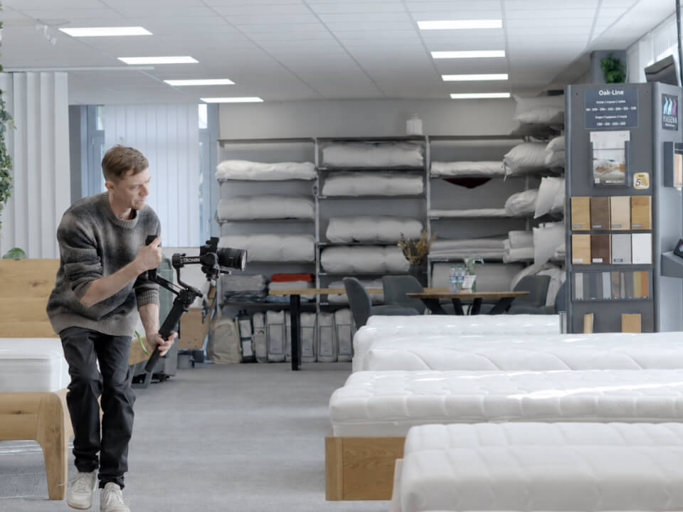 Mann filmt mit Kamera-Gimbal in einem Matratzen-Showroom mit vollen Regalen.