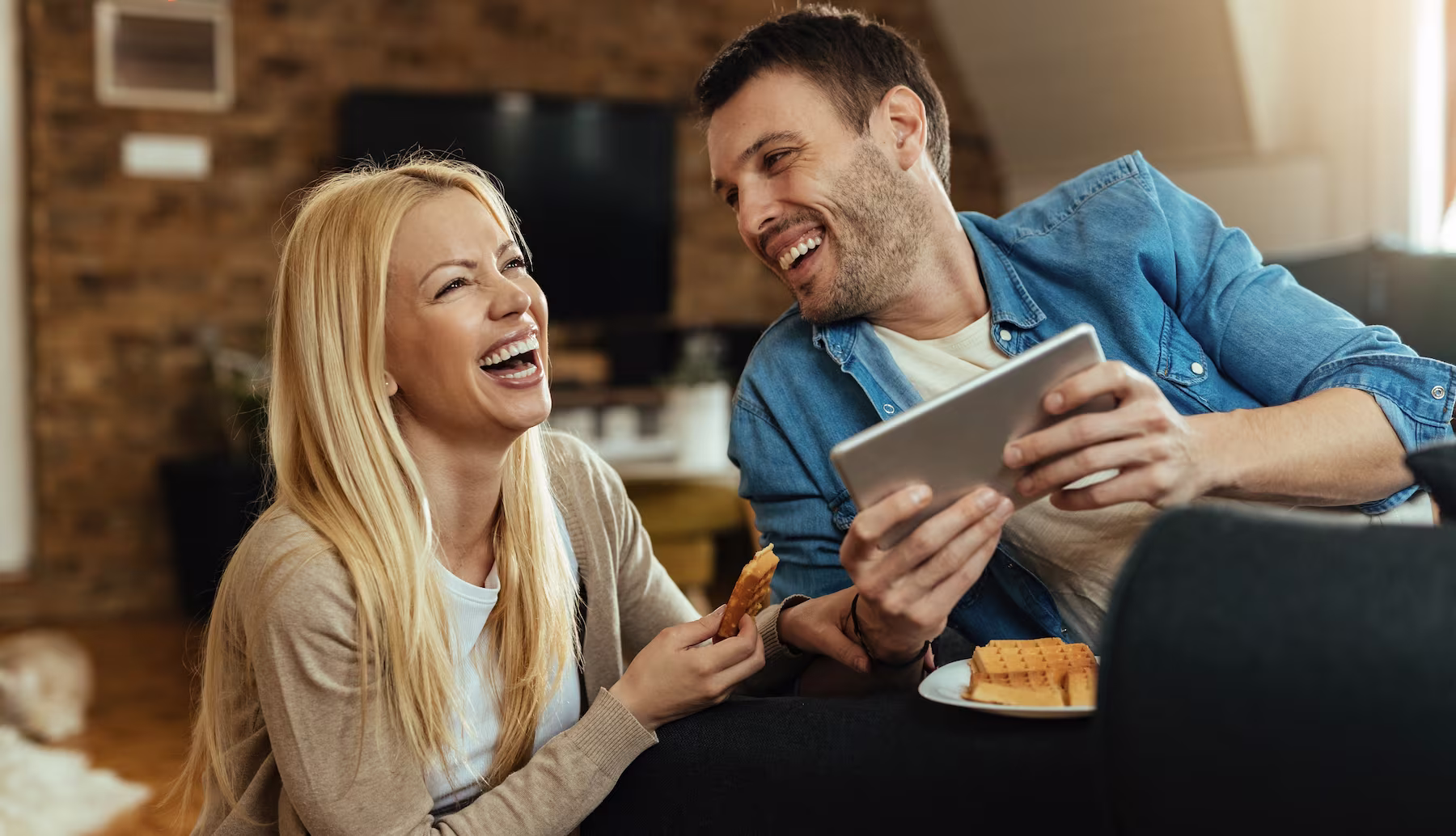 A candid photo of two young people laughing together against a light wooden wall background. One person has natural curly black hair and wears a white cable knit sweater, while the other has wavy brown hair and wears a gray sweater. Both appear to be sharing a genuine moment of joy and amusement.