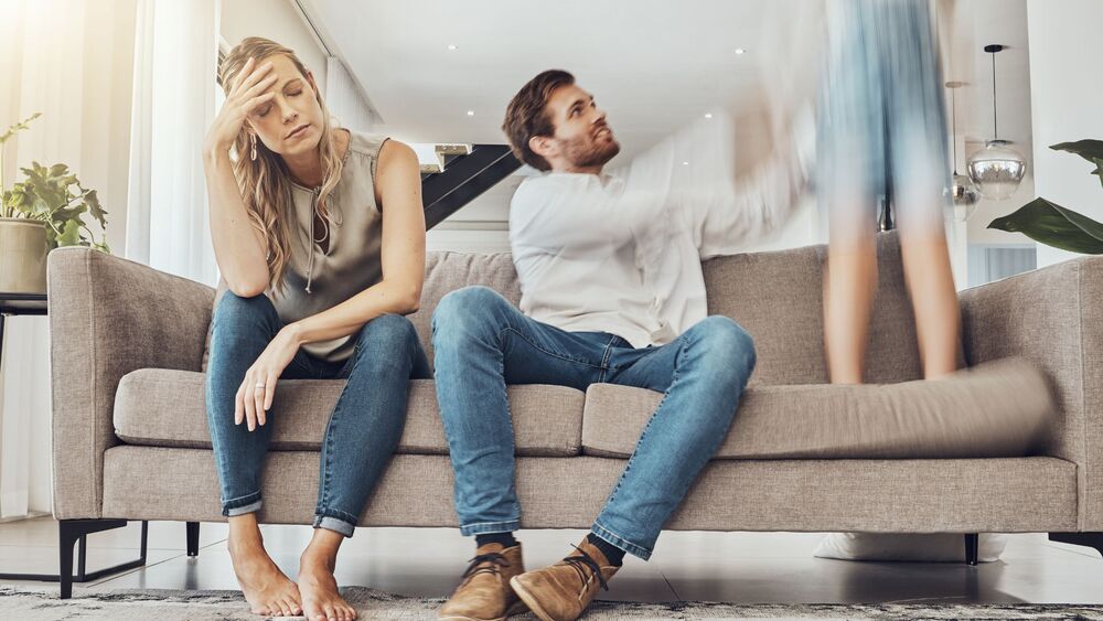 Tired couple sitting on couch while child jumps, symbolizing burnout and family stress.