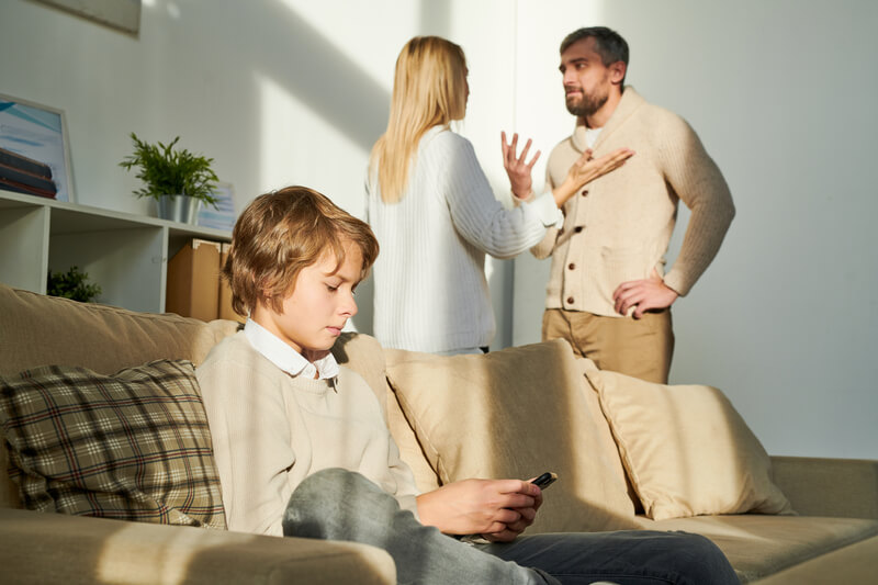 Child sits closed-off while parents argue in the background.