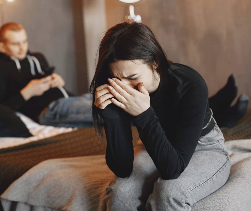 Sad woman crying while partner looks away at phone, showing distance and a lack of understanding.