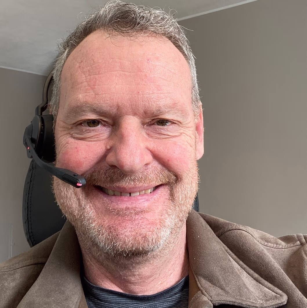 Lächelnder Mann mittleren Alters mit grauem Bart trägt Headset und braunen Mantel vor grauer Wand.