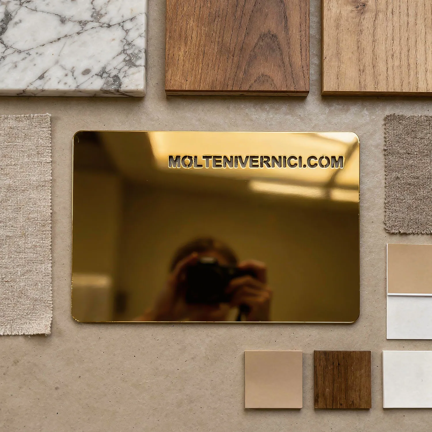 Campione di finiture con lastre in marmo, legno, tessuto e una lastra metallica riflettente dorata con la scritta moltenivernici.com.