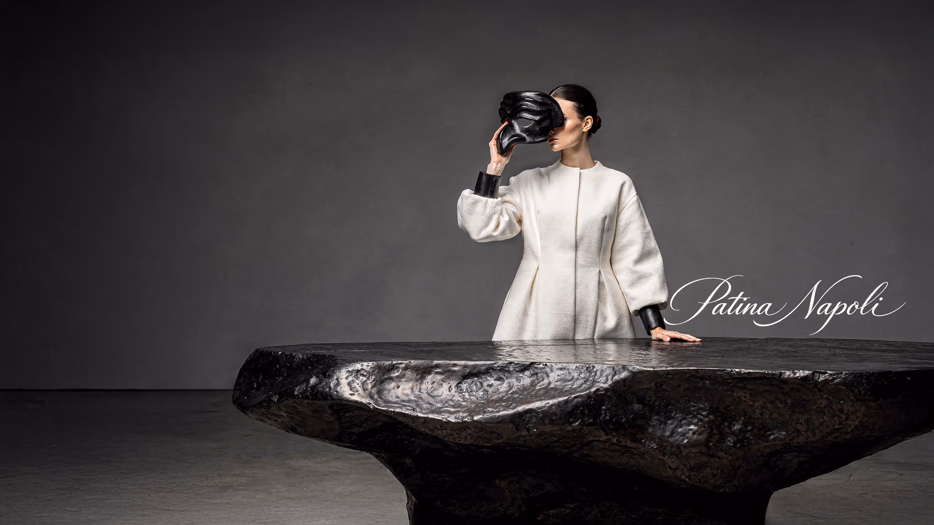 Luxury fashion editorial scene featuring a sculptural hammered black iron monolithic table in the foreground, with an Italian woman in an elegant ivory couture outfit standing behind it, gently removing a traditional black Pulcinella mask; minimalist architectural backdrop with soft natural daylight and “Patina Napoli” script on the wall.
