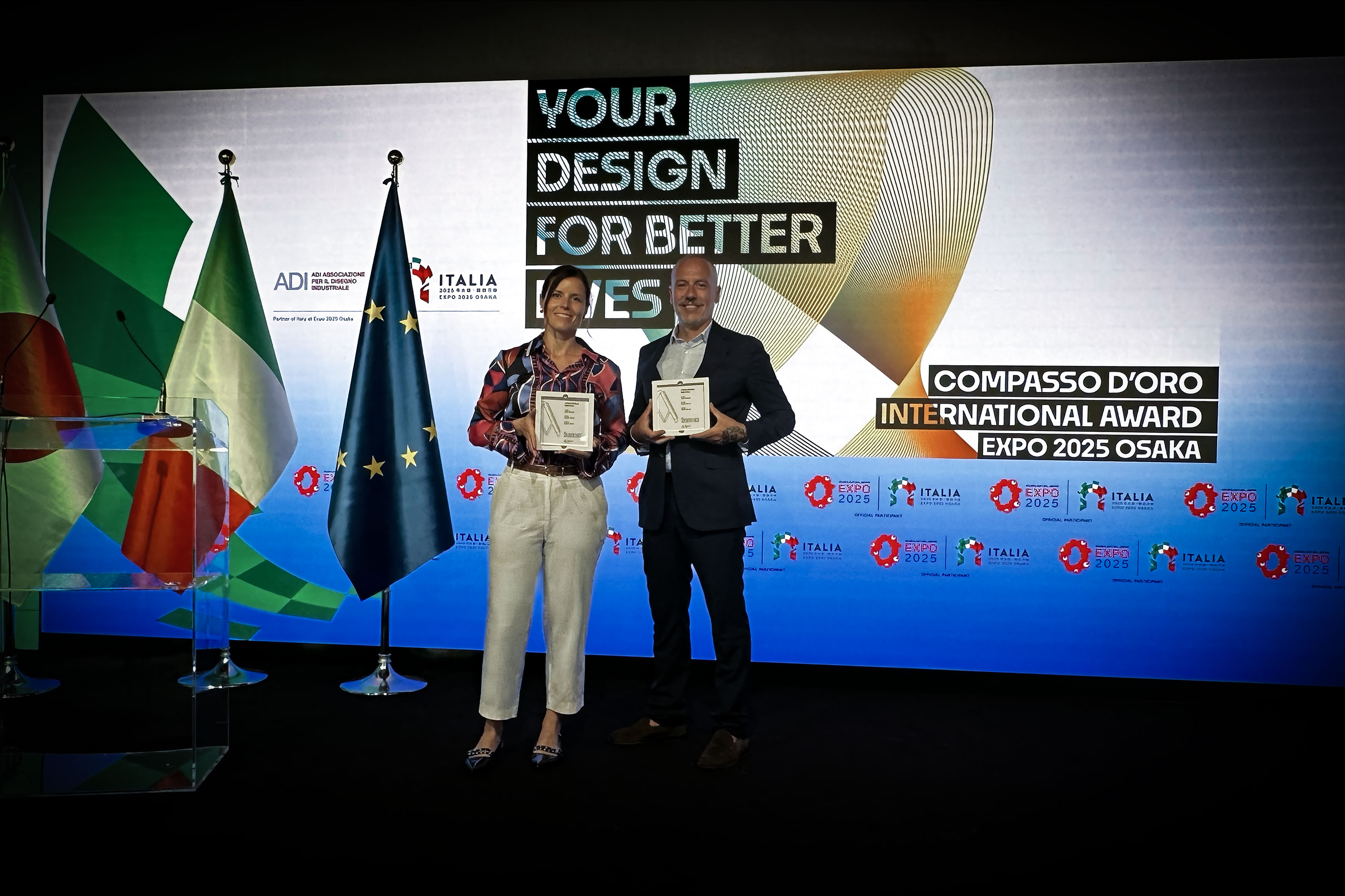 Two award recipients posing with Compasso d'Oro awards at Expo 2025 event