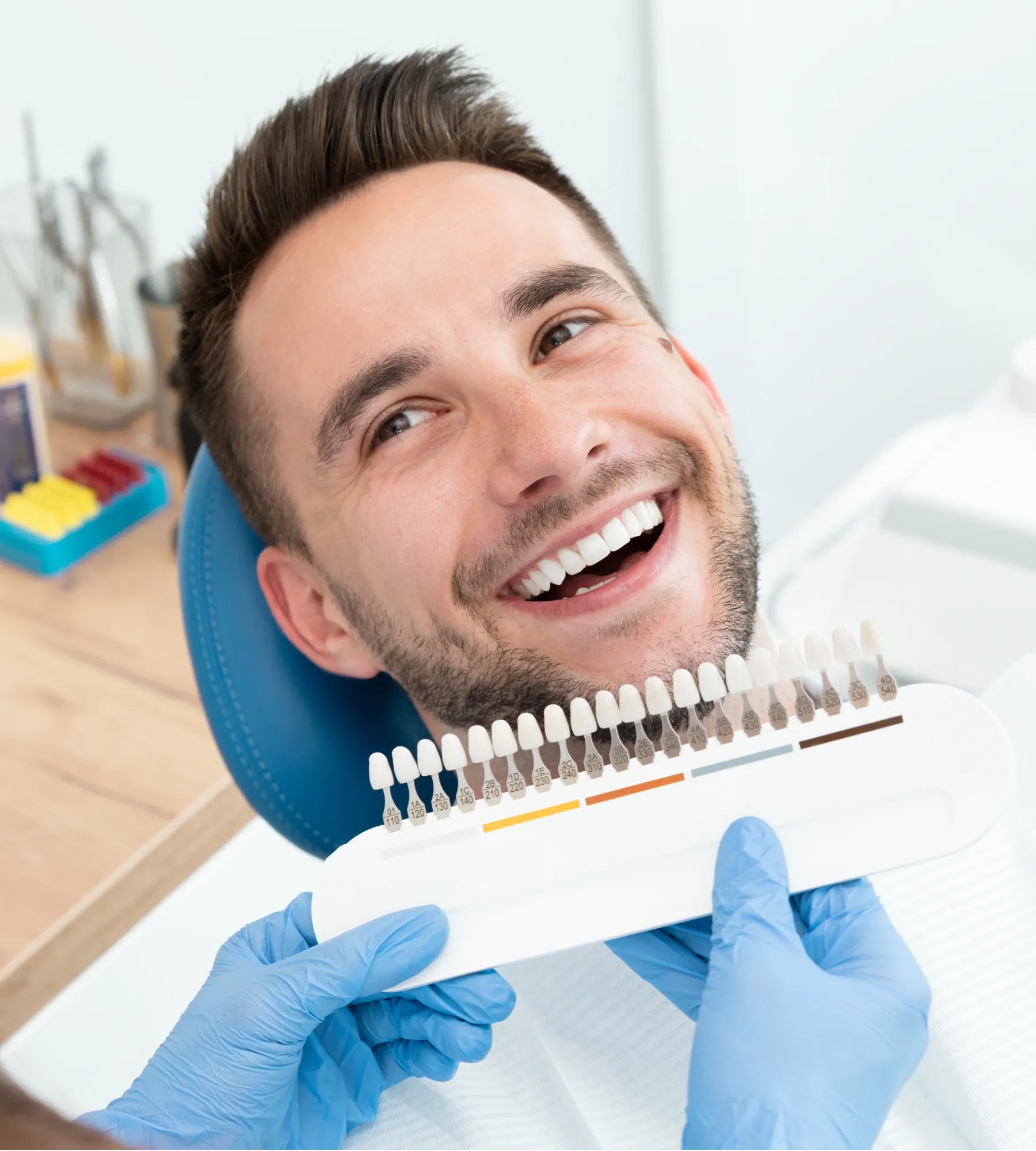 Smiling man in dental chair with a dental shade guide held against his teeth by gloved hands.