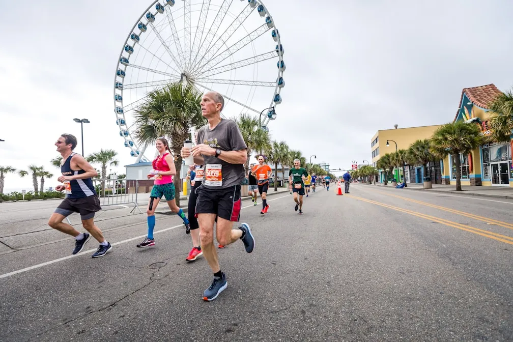 Runners participating in a road race near a large Ferris wheel on an overcast day. They are on a street lined with palm trees and colorful buildings. Some wear bib numbers and athletic gear.