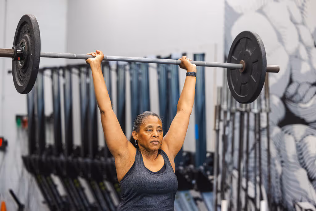 Middle-aged woman lifting a barbell overhead in a gym with workout equipment in the background.