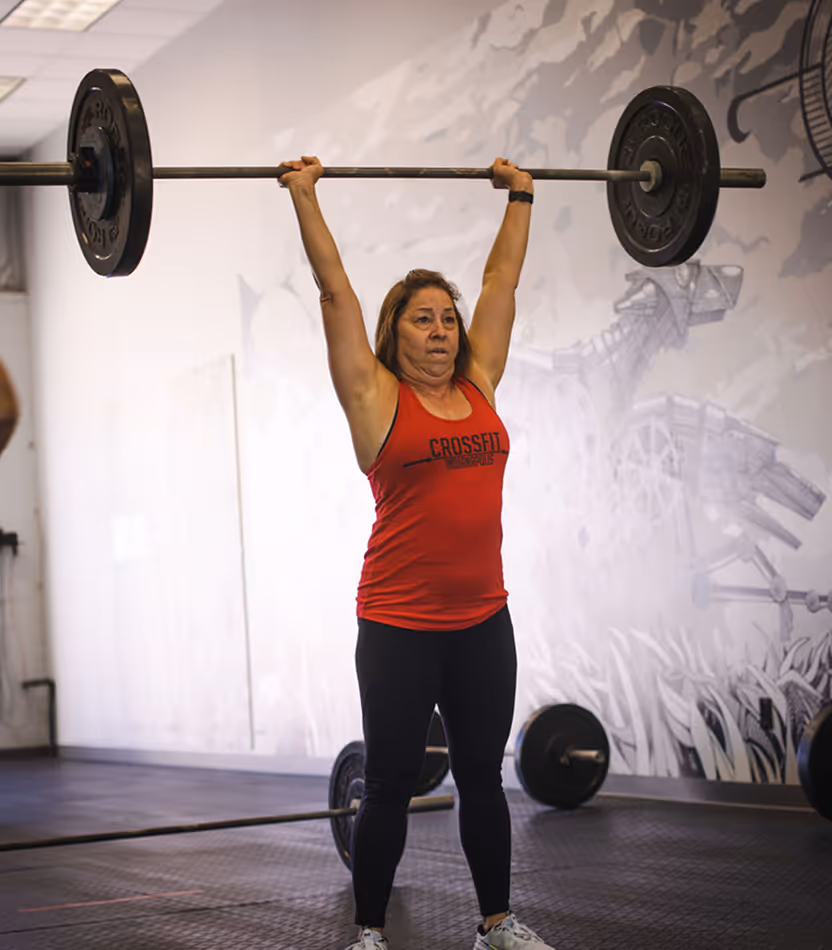 Woman in red CrossFit tank top lifting a weighted barbell overhead in a gym.