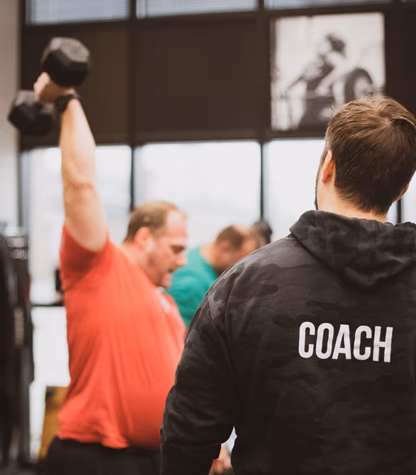 A coach in a black hoodie watches a man in a red shirt lifting a dumbbell overhead in a gym.