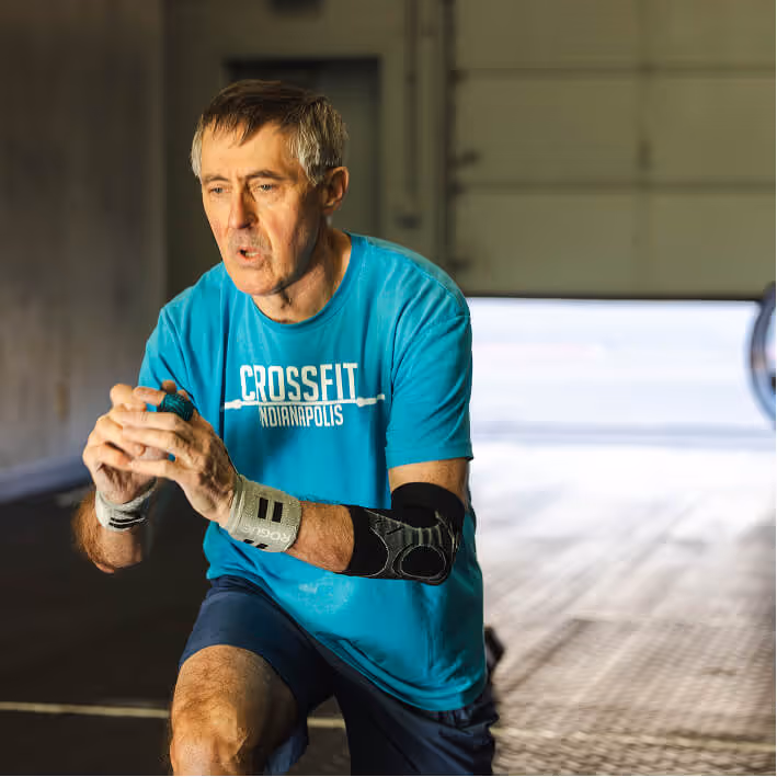 Middle-aged man wearing a turquoise CrossFit Indianapolis shirt and protective gear performing a lunge exercise indoors.