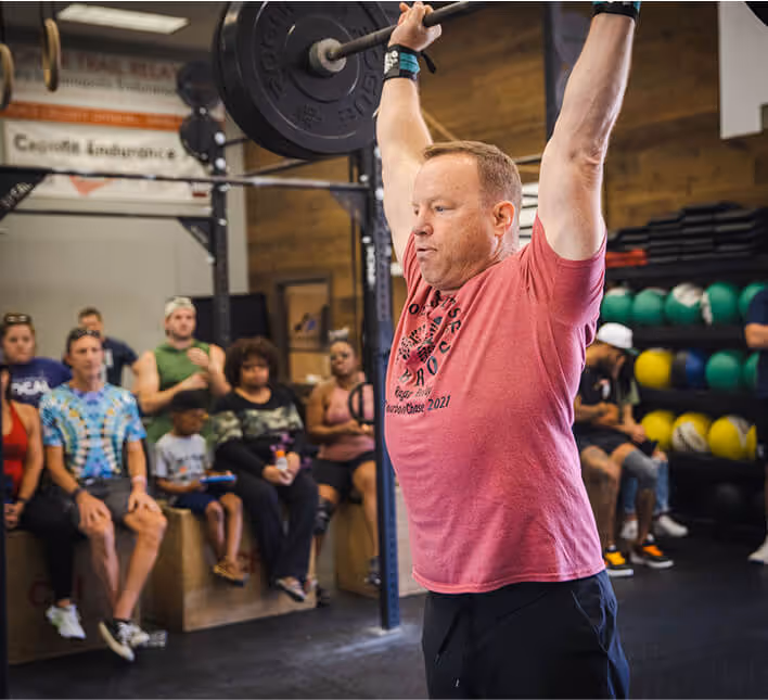 Man lifting a heavy barbell overhead in a gym while others watch and sit nearby.