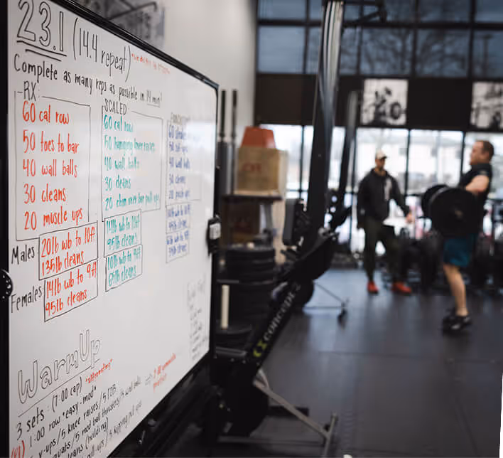 Whiteboard with CrossFit workout instructions for 23.1 including exercises like calorie row, toes to bar, wall balls, cleans, and muscle ups, with a rowing machine and two people exercising in a gym.