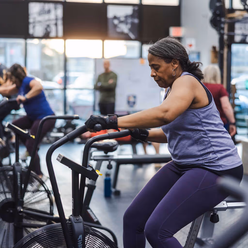 Mature woman in athletic wear exercising on a stationary bike in a gym with other people in the background.