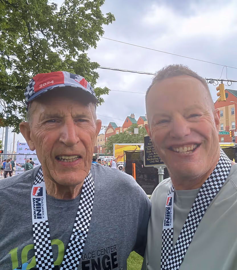 Two men with checkered race medals around their necks smiling outdoors at a race event.