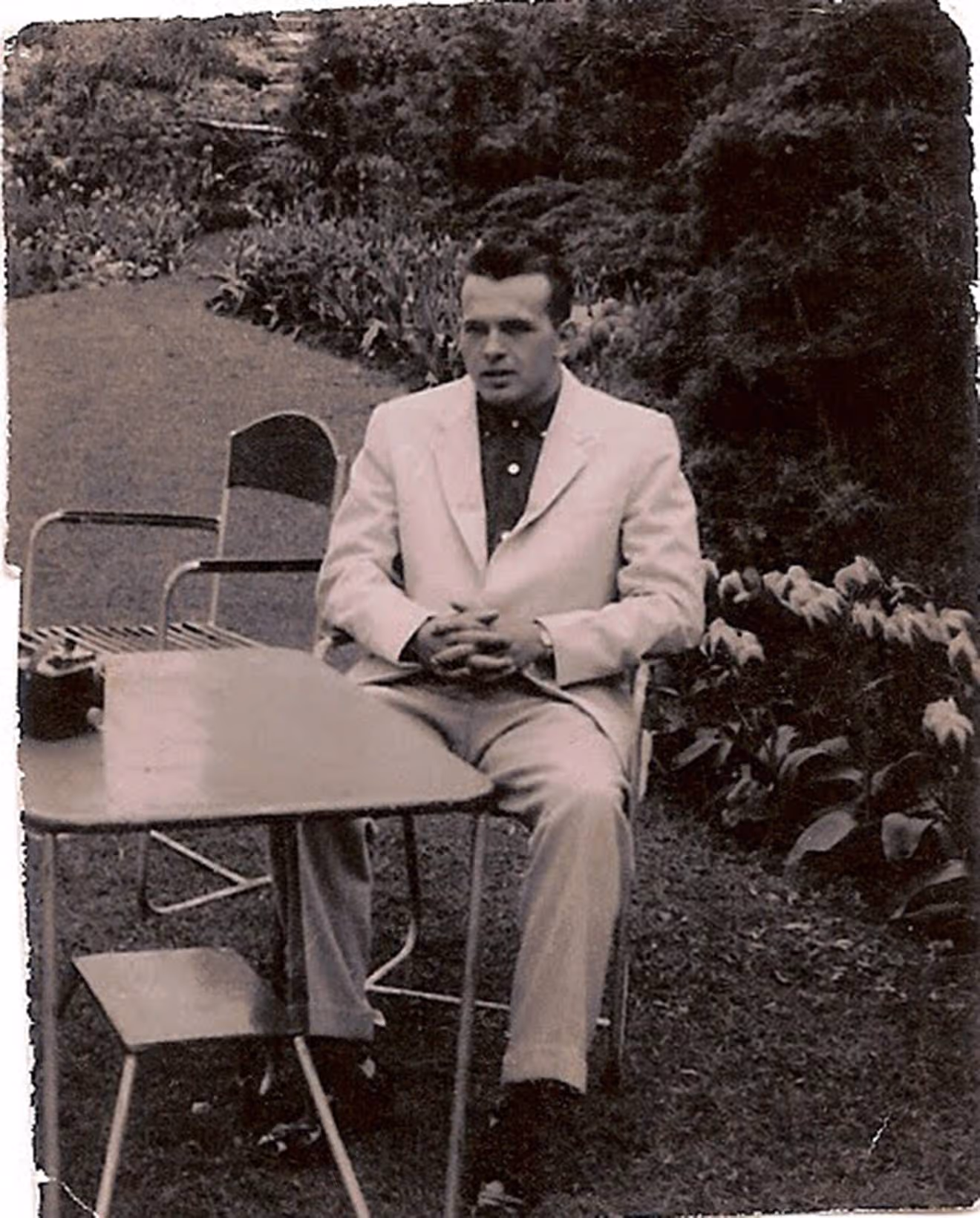 Stephen's father, Bob, dressed in a white suit, sits at a table outside with his hands clasped in his lap. Behind him are trees and blooming flowers.