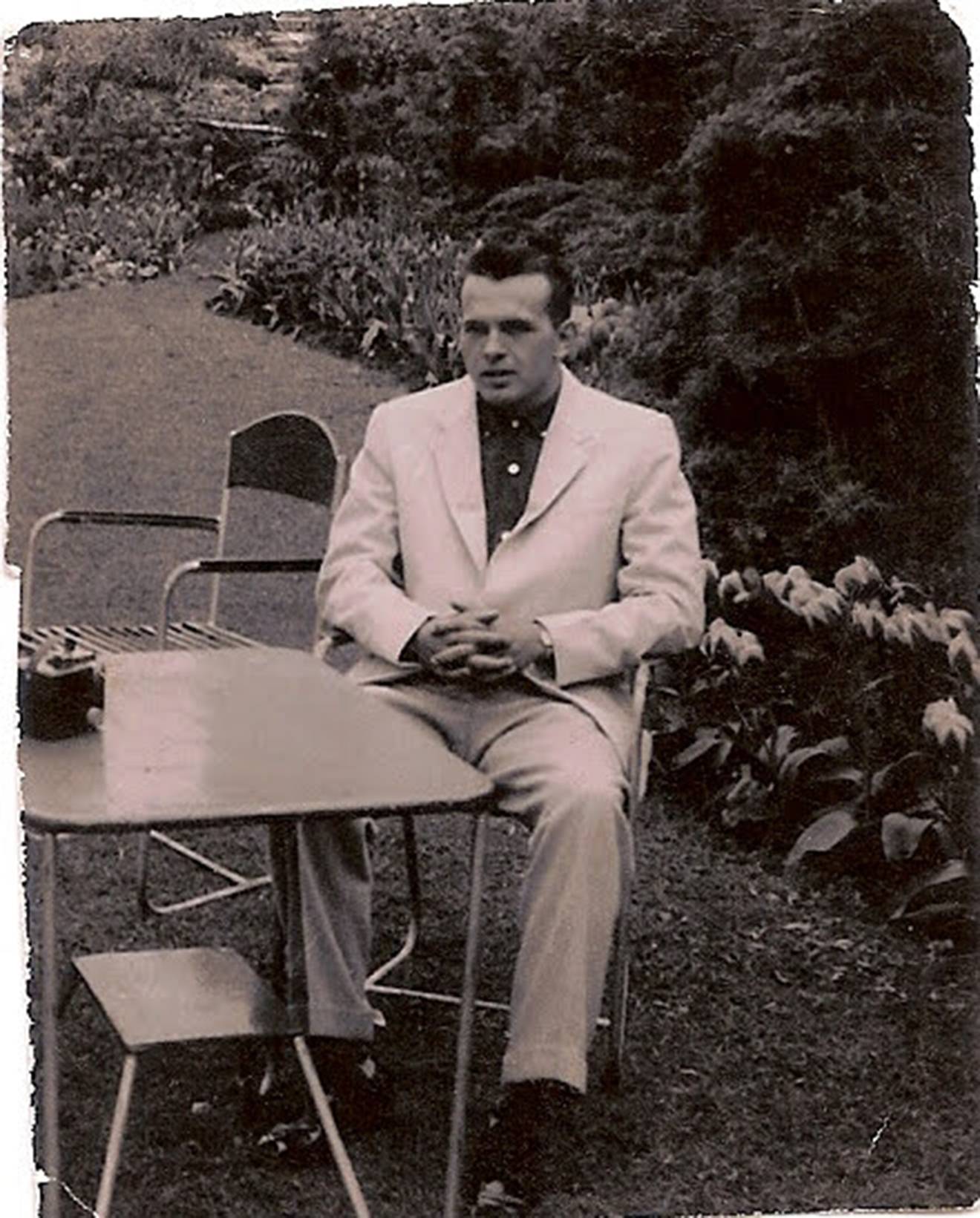 Stephen's father, Bob, dressed in a white suit, sits at a table outside with his hands clasped in his lap. Behind him are trees and blooming flowers.