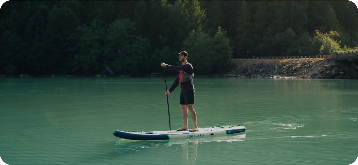 Man paddleboarding alone on a turquoise lake surrounded by forested hills.