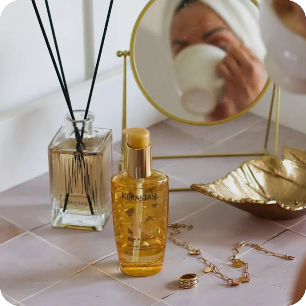Kerastase hair oil bottle on a bathroom counter beside a reed diffuser, gold tray, and mirror.