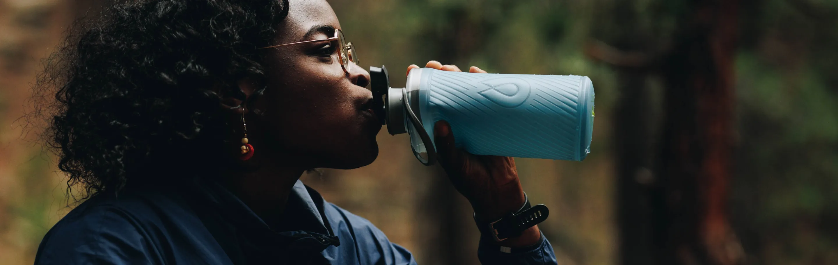 Woman drinking from a light blue insulated water bottle in a forest setting.
