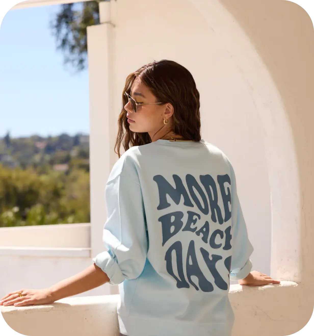 Woman wearing a light blue Vintage Havana sweatshirt printed with 'More Beach Days' on a sunny balcony.