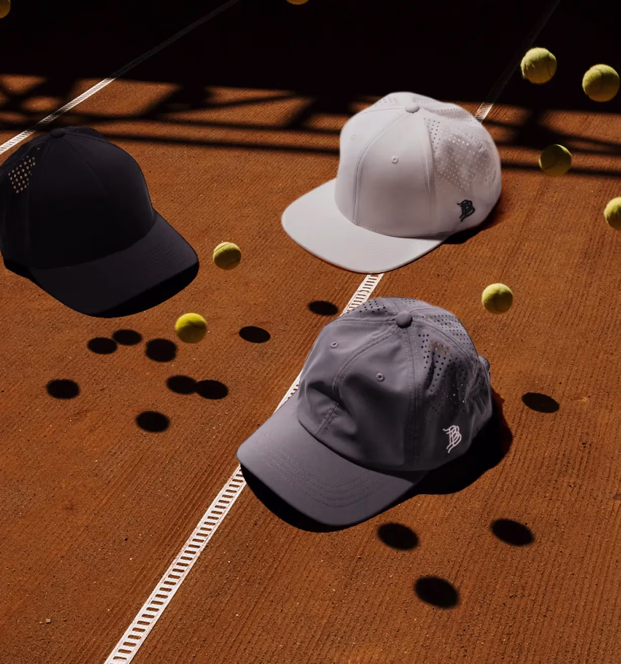 Three Branded Bills baseball caps displayed on a clay tennis court surrounded by tennis balls.