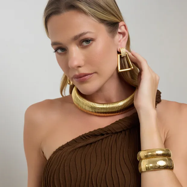 Model wearing gold statement jewelry including a chunky choker necklace, cuff bracelets, and drop earrings.