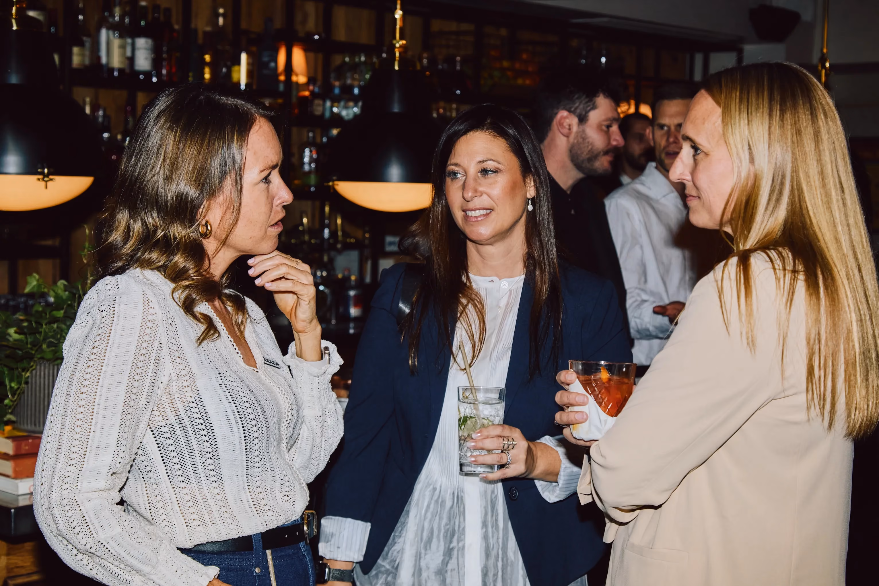 Three attendees chatting with drinks in hand near the bar at an ehouse networking event.