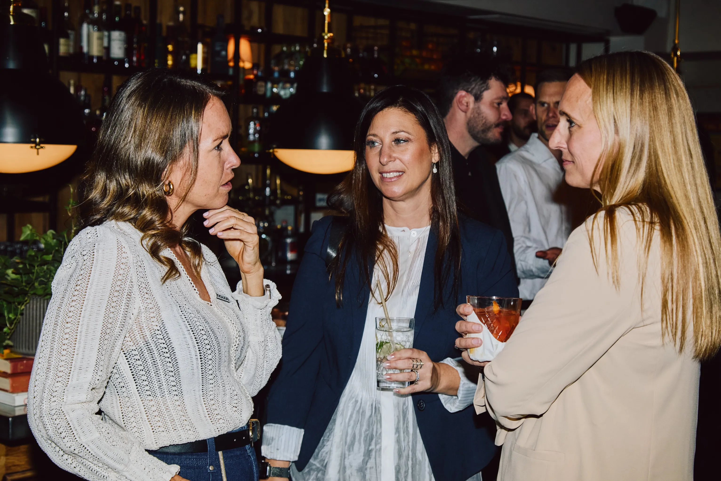 Three attendees chatting with drinks in hand near the bar at an ehouse networking event.