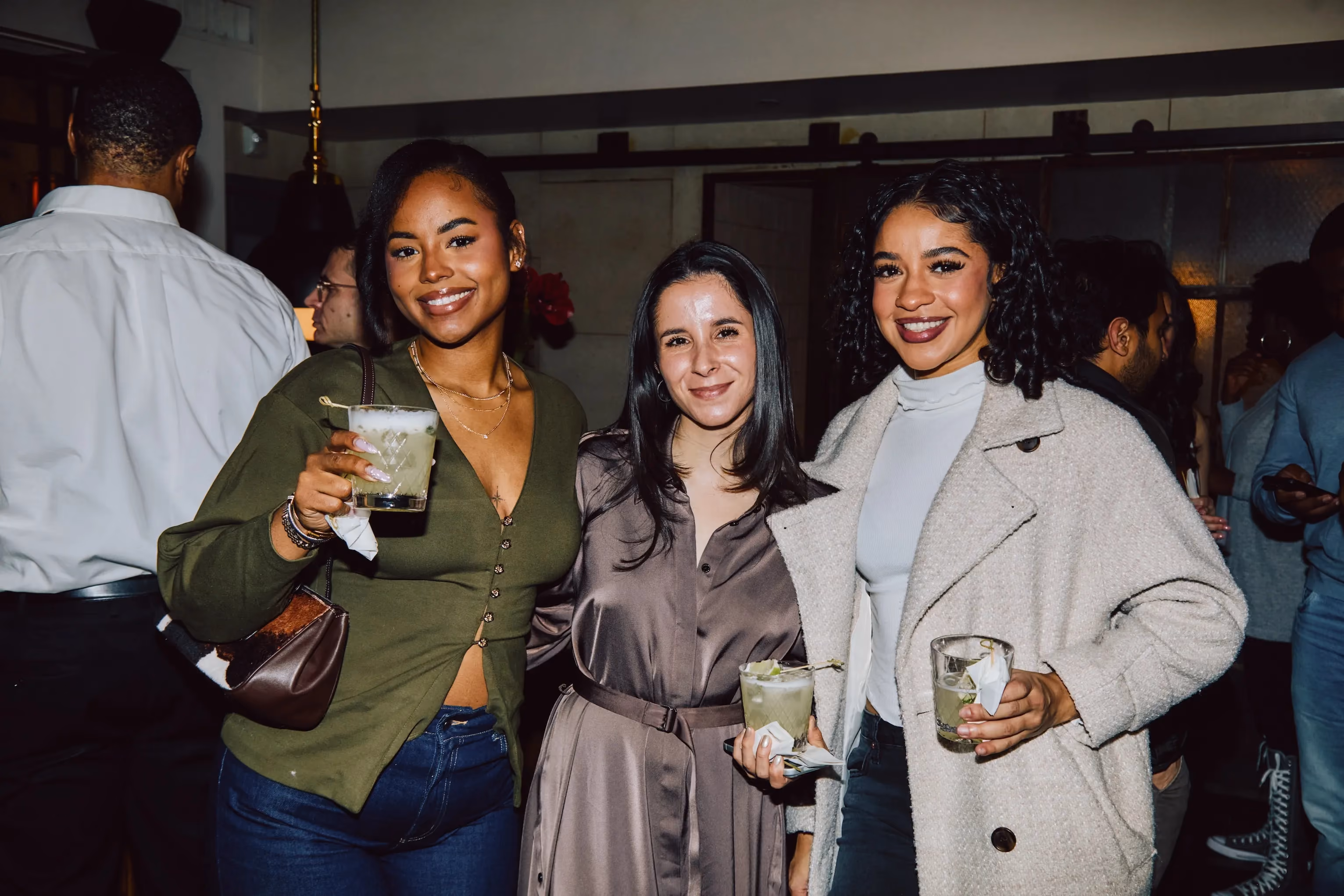 Three women holding cocktails and smiling at an evening ehouse networking event.