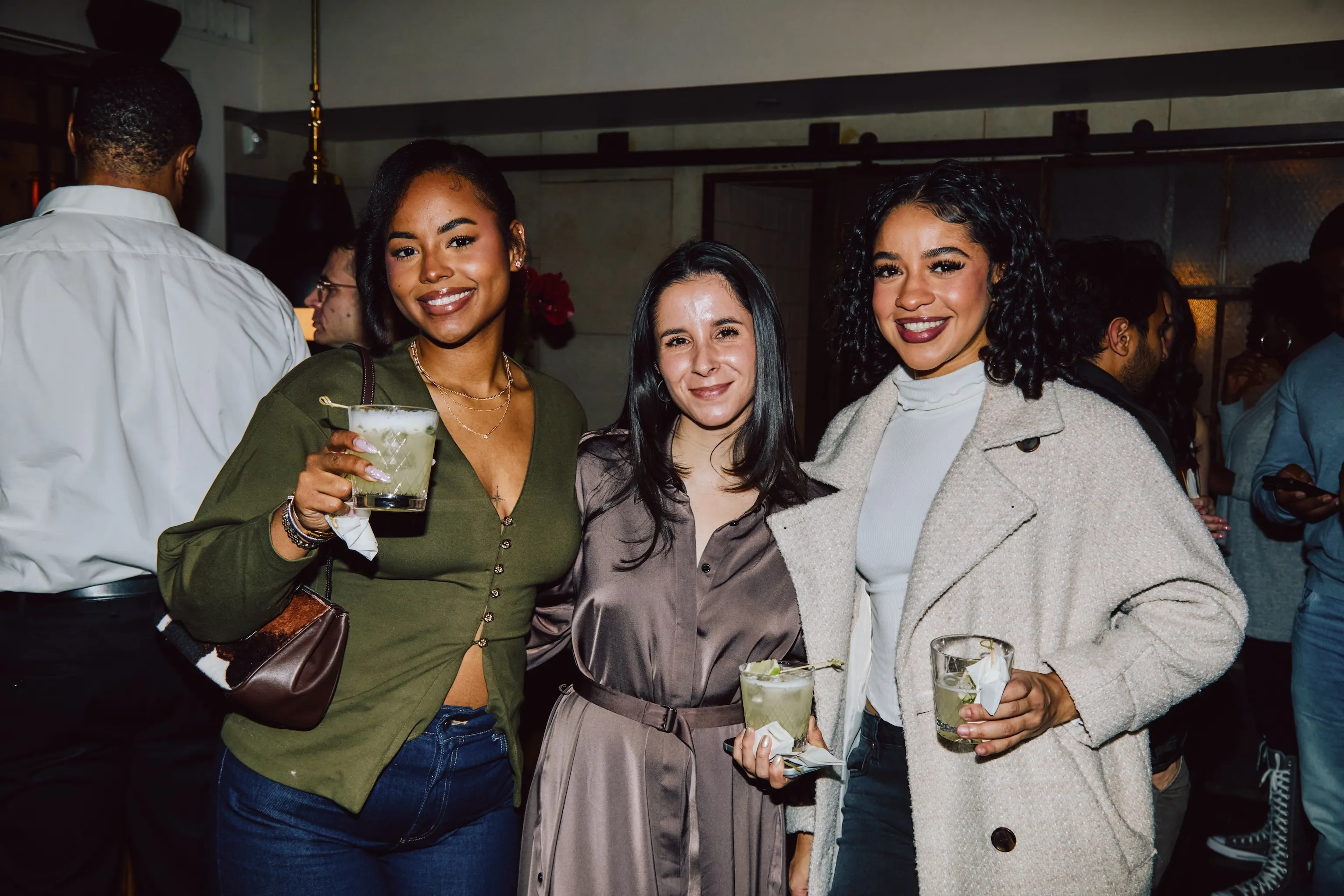 Three women holding cocktails and smiling at an evening ehouse networking event.