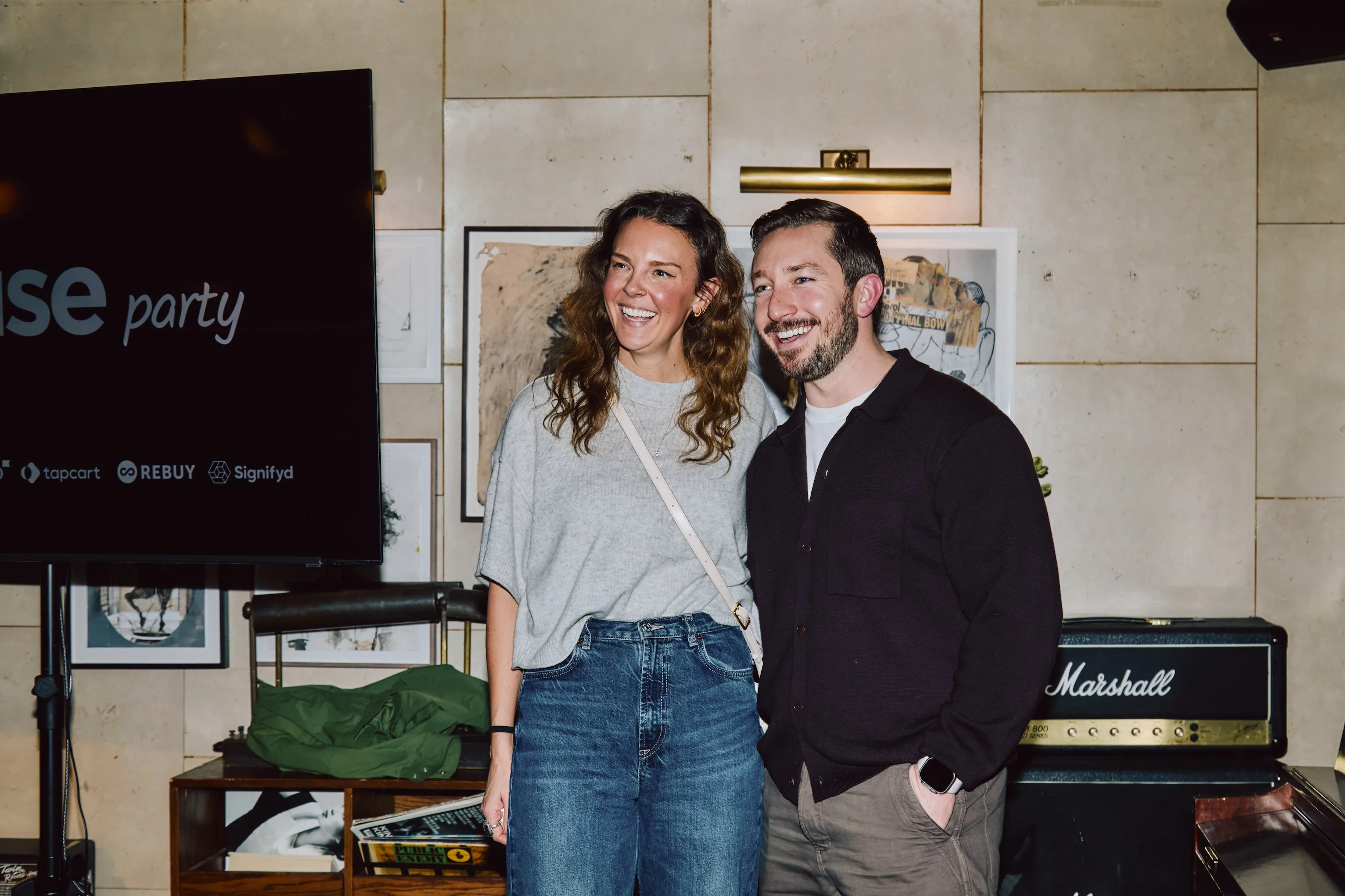 Two colleagues smiling at an ehouse party event with a screen showing sponsor logos Topcart, Rebuy, and Signifyd.