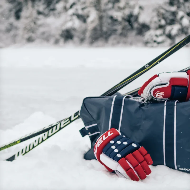 Winnwell hockey sticks, gloves, and equipment bag resting in the snow beside a frozen pond.